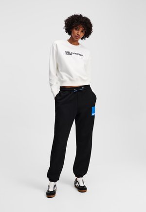 White sweatshirt with black "KARL LAGERFELD JEANS" text, paired with black sweatpants featuring a blue patch and drawstring, and black sneakers.