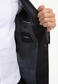 Wnętrze czarnej kurtki, z gładką szarą podszewką, kieszenią z metką i jednym zapięciem na guzik. Zewnętrzna tkanina wydaje się elegancka.