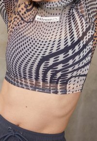 Průhledný cropped top v šedé barvě s abstraktními vzory v černé a hnědé, s kulatým výstřihem a elastickým lemem. Viditelný štítek značky.