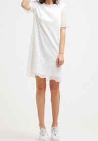 Valkoinen pitsimekko, jossa on lyhyet hihat ja aallotettu helma. Mekossa on kukkakuvio ja kevyt materiaali, ja se on yhdistetty valkoisiin tennareihin.