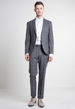 Grau karierter Anzug mit tailliertem Blazer und schmal zulaufenden Hosen, kombiniert mit einem weißen Hemd und hellgrauen Schuhen.