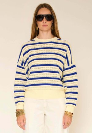 Femme portant des lunettes de soleil oversize, un pull tricoté à rayures bleues et crème, un pantalon blanc, des boucles d'oreilles en or, des bracelets et des bagues, debout droit.