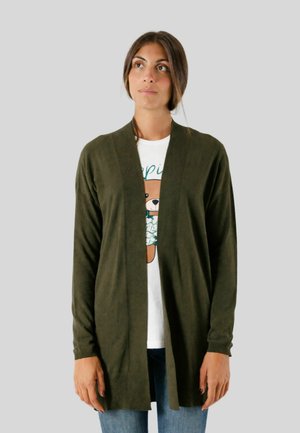 Donna che indossa un cardigan verde scuro aperto sopra una T-shirt bianca con una grafica di un orsetto, jeans blu, in piedi contro uno sfondo semplice.