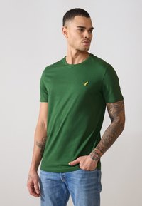 Grønn kortermet t-skjorte i et glatt stoff med rund hals, med en liten gul logo på venstre bryst. Kombinert med blå jeans.