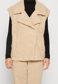 Beige ärmellose Sherpa-Weste mit breiten Revers und asymmetrischem Reißverschluss, getragen über einem schwarzen gerippten Rollkragenpullover und beigen Hosen.
