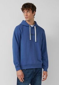 Giovane uomo che indossa una felpa con cappuccio blu e lacci bianchi, con jeans blu scuro, che guarda di lato su uno sfondo semplice.