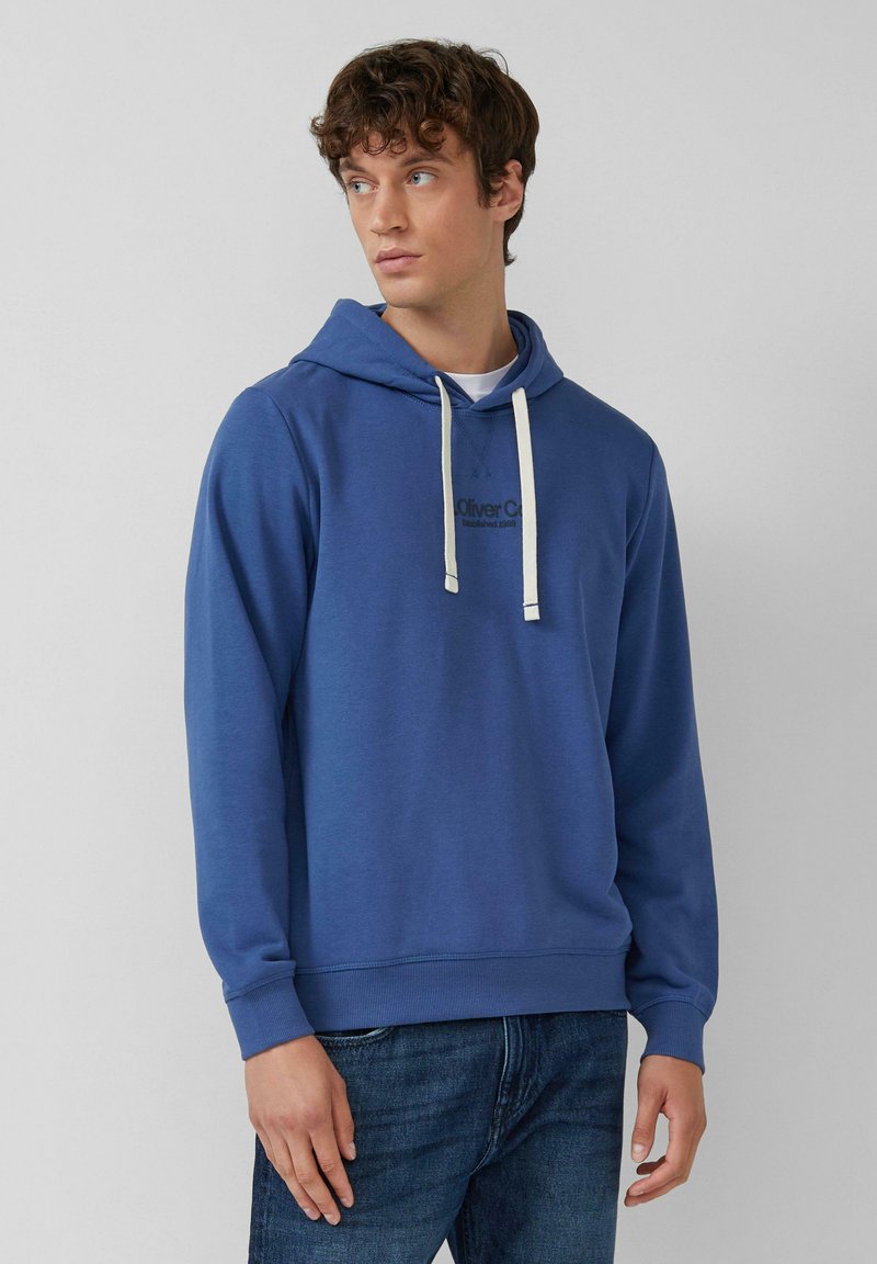 Giovane uomo che indossa una felpa con cappuccio blu e lacci bianchi, con jeans blu scuro, che guarda di lato su uno sfondo semplice.