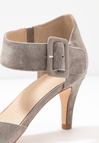 Harmaa mokkanahkainen korkea kenkä nilkkahihnalla, jossa on suorakulmainen solki. Suora nahkasisus ja hoikka, kapeneva korko.