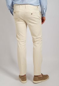 Beige Baumwollhosen mit schmaler Passform, ausgestattet mit Rückentaschen mit Knöpfen, kombiniert mit hellbraunen Slippern.