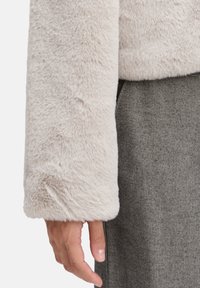 Lys beige faux fur jakkeærme med en blød tekstur; parret med et gråt herringbone-stof nedenunder. Hånd hviler let på beklædningen.