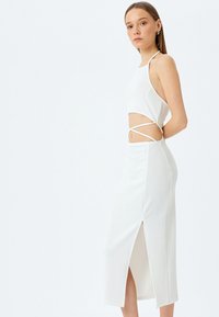 Weißes geripptes Kleid mit einer figurbetonten Passform, ausgeschnittenem Mittelteil und seitlichem Schlitz. Details umfassen einen Halterneck und kreuzweise Akzente an der Taille.