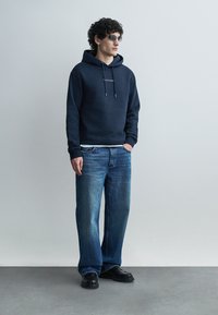 Muž s kučeravými vlasmi, ktorý má na sebe tmavé slnečné okuliare, tmavomodrú mikinu s kapucňou, voľné modré džínsy a čierne topánky, stojí s jednou rukou v vrecku.