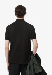 Camisa polo negra de manga corta, textura suave, cuello clásico. Modelo sostiene una chaqueta verde oscura, visto desde la espalda sobre un fondo blanco.