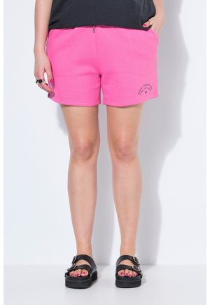 Persona che indossa pantaloncini rosa acceso con la scritta "Loud Proud and Full of Love", sandali neri e top nero, in piedi su uno sfondo bianco.