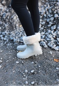 Bottes en caoutchouc gris clair avec un revers intérieur crème et duveteux, présentant une semelle texturée et un bout arrondi, posées sur un terrain gravillonné.