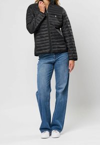 Černá prošívaná bunda s předním zipem, proužkovaným vzorem prošití a malým logem na hrudi. Kombinována se světle modrými džíny širokých nohavic a bílými botami.