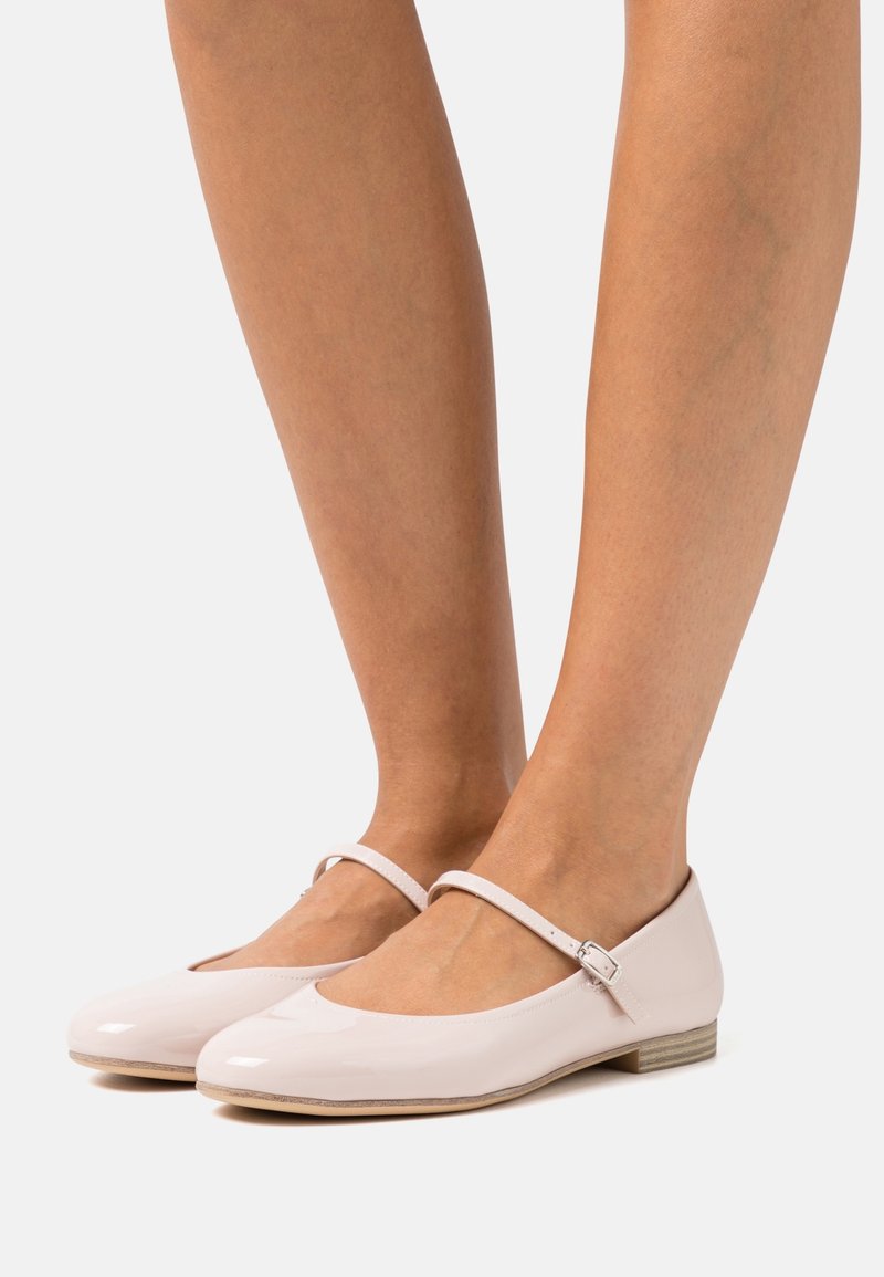 Ballerines Mary Jane rose pâle brillantes avec une fine sangle à boucle portées pieds nus sur un fond blanc.