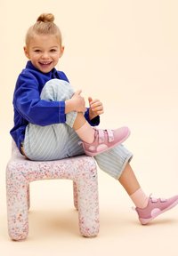 Blauwe jas, lichtgestreepte broek en roze sneakers met klittenband. Kind zit op een gespikkelde witte stoel tegen een zachte pastelachtergrond.