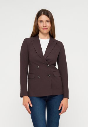 Femme aux cheveux bruns et raides portant un blazer marron foncé à double boutonnage sur un haut blanc et un jean bleu, debout devant un fond uni.