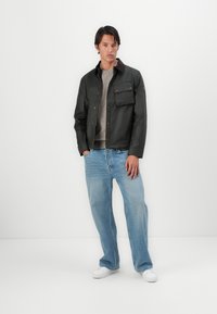 Zwarte gewaxte jas met een bruine kraag, twee voorkruiden, grijs sweater eronder, blauwe spijkerbroek en witte sneakers. Model staat tegen een witte achtergrond.