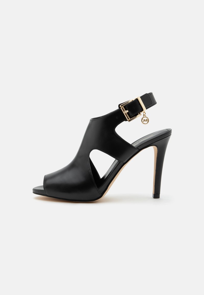 Sandale noire en cuir à talon haut avec bout ouvert, découpes latérales, bride à la cheville, boucle dorée et petit pendentif rond sur un fond clair.