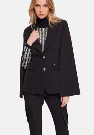 Blazer nero con design a mantella, con bottoni dorati e due tasche frontali. Indossato sopra una maglia nera e bianca a motivo.