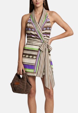 Donna che indossa un abito avvolgente senza maniche con scollo a V e motivi geometrici in marrone, verde e viola, che tiene una borsa intrecciata marrone.