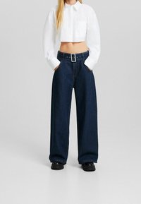 Vit kortärmad skjorta med krage, i kombination med vida mörkblå jeans med bälte och silverspänne, samt svarta platåskor.