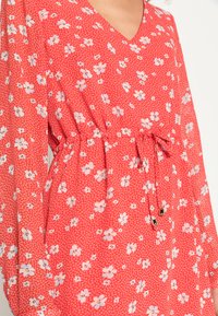 Vestido floral vermelho em tecido leve, com decote em V, mangas longas, cintura ajustada e detalhes de cordão com acabamentos em dourado.