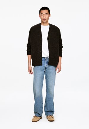 Mężczyzna w ciemnobrązowym kardiganie na guziki, białej koszulce, jasnoniebieskich dżinsach i beżowych butach, stojący na tle białego tła.