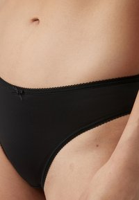 Gros plan d'une personne portant un sous-vêtement noir uni de style bikini avec un petit nœud décoratif sur la ceinture avant.