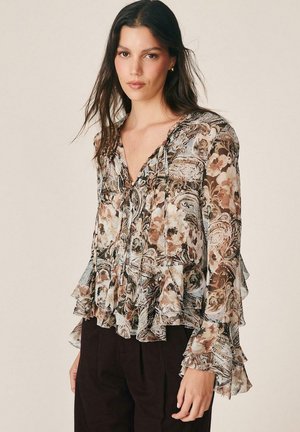 Mujer con cabello largo y oscuro lleva una blusa transparente con estampado floral paisley, con puños y bajo con volantes, combinada con pantalones oscuros, de pie frente a un fondo liso.