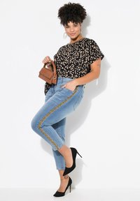 Jeans de mezclilla azul claro con franjas laterales metálicas doradas, combinados con una blusa de estampado de leopardo en negro y beige y tacones altos negros. Se sostiene un pequeño bolso marrón.