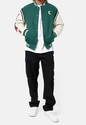 Veste de varsity verte et crème avec des boutons pression, des poignets rayés et un logo sur la poitrine, associée à un pantalon cargo noir et des baskets blanches.