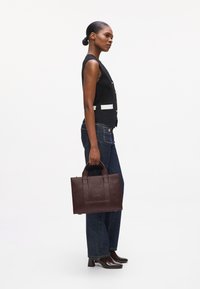 Kvinne iført svart vest og mørke jeans holder en brun skinnveske, stående i profil med håret i en lav hestehale og iført høye sko.