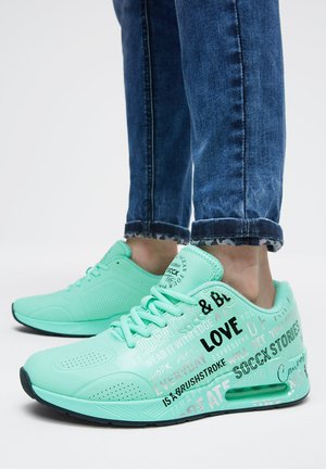 Person wearing light turquoise sneakers with black and white text designs, paired with rolled-up dark blue jeans, standing on a white background.