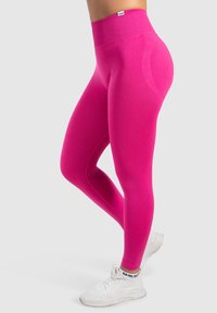 Legginsy z wysokim stanem w jaskrawym różu, charakteryzujące się gładkim, elastycznym materiałem i dopasowanym krojem, w parze z białymi butami sportowymi.