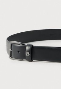 Schwarzer Ledergürtel mit silberner Metallschnalle, kleinem Logoakzent in der Nähe der Schnalle und verstellbaren Löchern entlang des Riemens zur Größenanpassung.