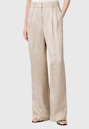 Pantalon beige taille haute à jambes larges avec plis devant, porté avec des sandales noires à bouts ouverts, mains dans les poches, sur fond gris clair.