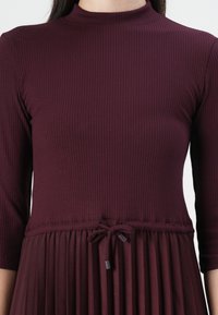 Burgunderkjole med ribbet topp og elastisk midje; plissert skjørt med knytedetalj. Lange ermer, høy hals, glatt tekstur.