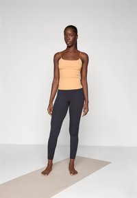 Tan scoop-neck tank top og mørkegrå leggings, tettsittende, ribbet tekstur, modell som står barbent på en lys beige matte mot en grå bakgrunn.