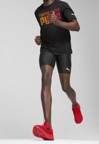 T-shirt de sport noire avec "FOREVER FASTER" en couleurs dégradées, associé à un short noir et à des chaussures de course rouges avec une semelle texturée.