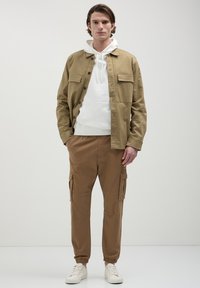 Homme portant une veste beige sur un hoodie blanc, un pantalon cargo marron et des baskets blanches, se tenant avec une main dans la poche devant un fond uni.