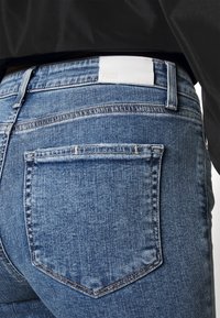 Jeans de denim azul con un diseño clásico de cinco bolsillos. En la parte trasera, se muestra una etiqueta de cuero blanco con texto en relieve. Se observa un ligero desvanecimiento.