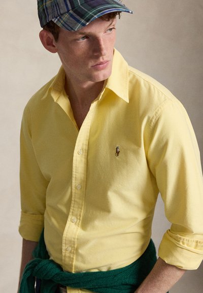 Mladý muž v žluté košili na knoflíky, modro-zelené kárované čepici a tmavě zeleném svetru uvázaném kolem pasu, který se dívá stranou.