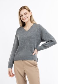 Szary sweter typu v-neck wykonany z miękkiego materiału, z długimi rękawami i prążkowanym wykończeniem. Noszony z beżowymi spodniami, podkreślającymi luźny krój.