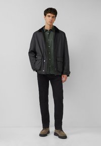 Veste légère noire à fermeture par boutons, deux poches avant, portée sur une chemise verte à motifs, associée à un pantalon noir et des chaussures marron.
