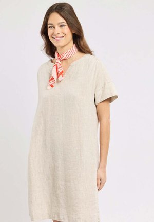 Femme portant une robe beige à manches courtes et un foulard rayé rouge et blanc autour du cou, souriante et regardant sur le côté devant un fond uni.