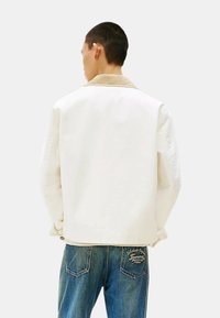 Giacca bianca con collo in corda color beige, caratterizzata da una vestibilità rilassata e spalle scese. Abbinata a jeans blu con dettagli del logo.