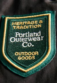 Zelená vyšívaná nášivka s nápisom "Dědictví a tradice", "Portland Outerwear Co." a "Outdoorové zboží", obšitá zlatou barvou na tmavém pozadí.